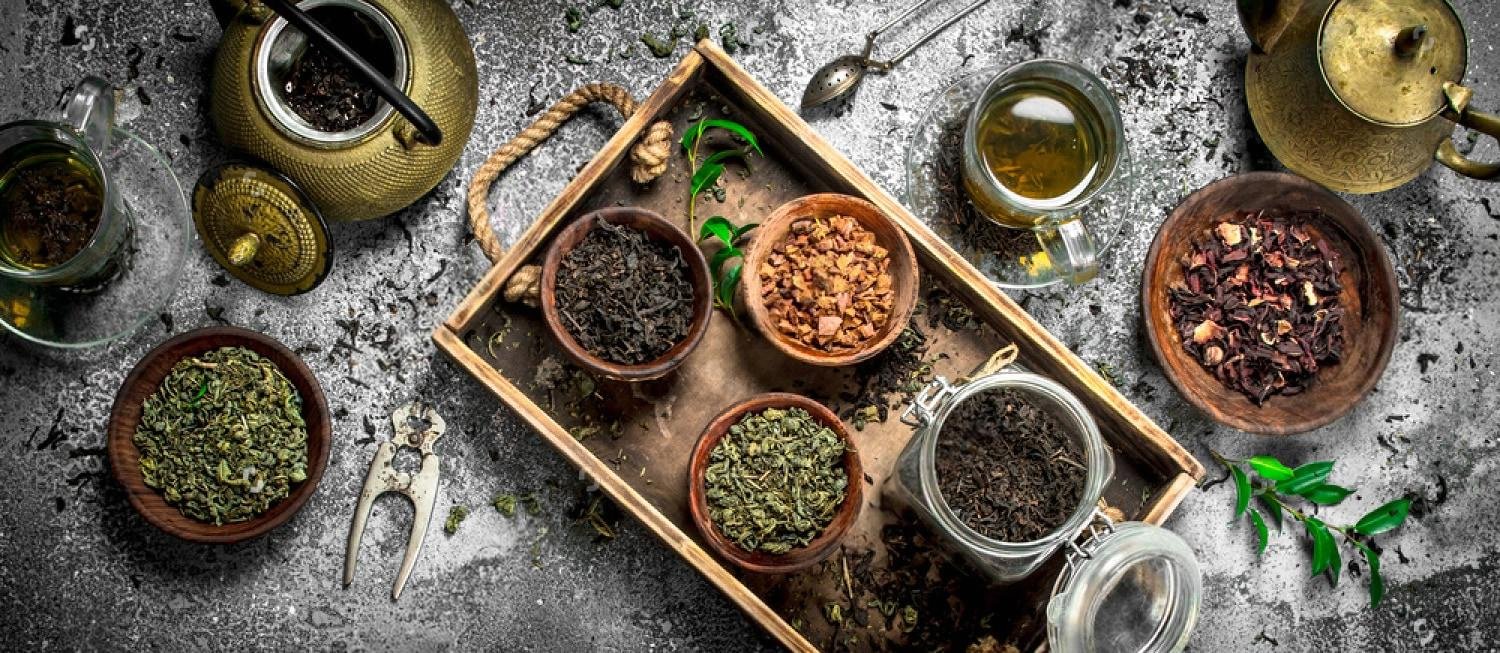 Wooden tray with bowls of green, black and dark Chinese teas beside brass teapots and glass cups